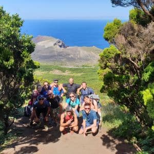 A WeRoad group trip poses on a dirt path framed by trees, overlooking a volcanic landscape and the ocean.