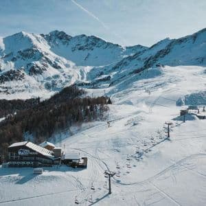 Veduta aerea di una stazione sciistica con rifugio e seggiovie su una montagna innevata sotto un cielo azzurro.