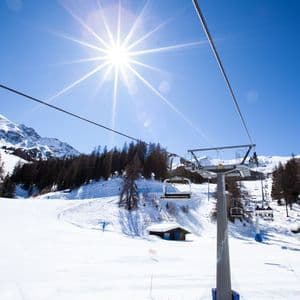 Una seggiovia si muove su un paesaggio montano innevato con pini sotto un sole splendente e un cielo azzurro limpido.