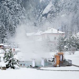 Persone si rilassano in una piscina termale all'aperto fumante, circondata da un paesaggio innevato con edifici e una fitta pineta.