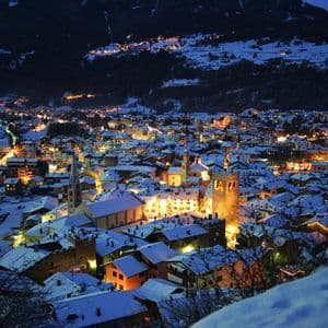Una città alpina con tetti innevati è illuminata a giorno di notte, incastonata ai piedi di una montagna scura.