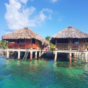 Due bungalow dal tetto in paglia, costruiti su palafitte sopra acque limpide color turchese, con una spiaggia sabbiosa e palme sullo sfondo.