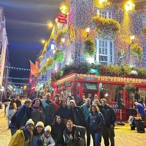 A WeRoad group trip smiling for a photo at night on a cobblestone street in front of a pub decorated with many fairy lights.