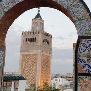 Un minarete de piedra con tejado verde, enmarcado por un arco ornamentado de azulejos coloridos, con vistas a un paisaje urbano al atardecer.