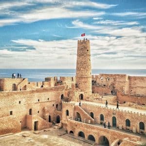 Un viaje en grupo de WeRoad explora una fortaleza histórica de piedra con una torre central y una bandera, con vistas al mar.
