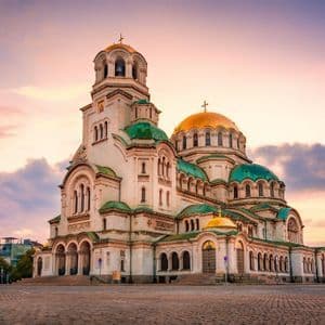 Una grande cattedrale di pietra bianca con cupole dorate e verdi si erge in una piazza di ciottoli al tramonto sotto un cielo colorato.