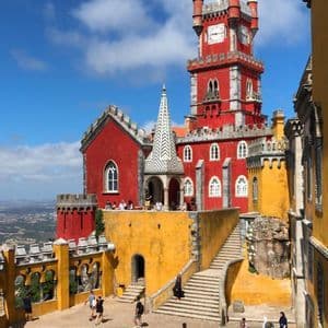 Menschen erkunden den Hof eines leuchtend rot-gelben Schlosses mit einem markanten Uhrturm unter einem teils bewölkten Himmel.