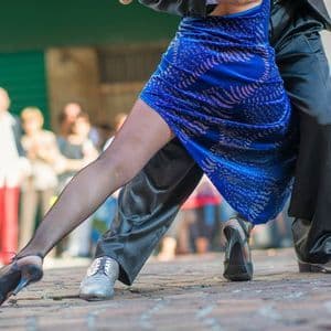 Un primer plano de las piernas de una pareja bailando tango en una calle adoquinada, con transeúntes difuminados al fondo.