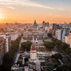 Una vista aerea di un denso paesaggio urbano incentrato su un grande edificio con una cupola verde e una piazza al tramonto.