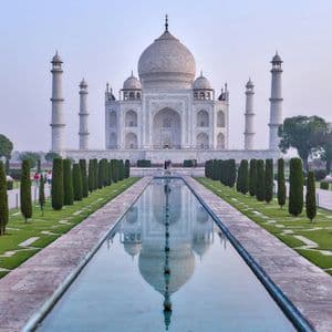 Un mausoleo di marmo bianco si riflette in una lunga vasca d'acqua, affiancato da giardini con cipressi sotto un cielo velato.