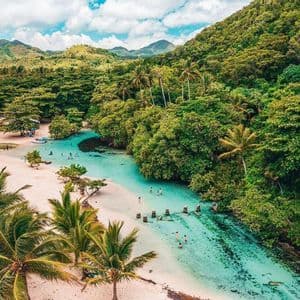 Vue aérienne de baigneurs dans les eaux turquoise limpides d'un lagon, bordé d'une plage de sable blanc et d'une forêt tropicale dense.