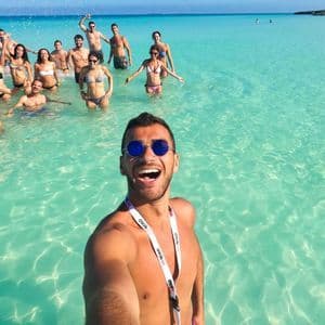 Ein Mann macht ein Selfie mit seiner WeRoad Gruppenreise, alle lächeln und stehen in klarem türkisfarbenem Wasser.