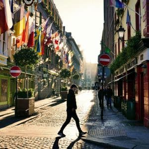 Une personne traverse une rue pavée ensoleillée, bordée de bâtiments ornés de nombreux drapeaux internationaux, et projetant une longue ombre.