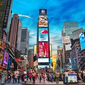 Una vista grandangolare di una piazza affollata con persone che camminano tra alti edifici coperti da grandi e luminosi cartelloni pubblicitari.