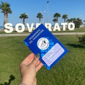Una mano regge un 'Passaporto del Viaggiatore' davanti a un grande cartello 'SOVERATO' su un prato con palme.
