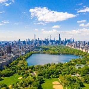 Vista aerea di un grande parco urbano con un lago centrale, delimitato da una fitta distesa di grattacieli sotto un cielo azzurro con nuvole.