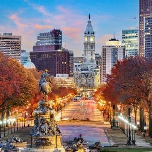 Una statua equestre si affaccia su un ampio viale alberato con alberi autunnali che conduce a uno skyline cittadino con una torre dell'orologio al tramonto.