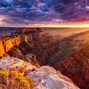 Un vasto canyon roccioso è illuminato dal sole al tramonto, con raggi di luce che filtrano attraverso nuvole viola drammatiche.