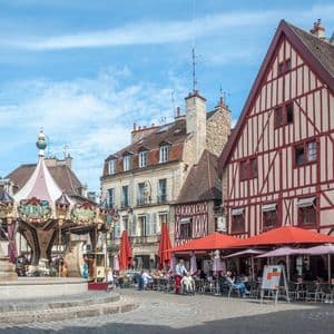 Ein europäischer Stadtplatz mit einem Brunnen, einem Karussell und einem Straßencafé neben traditionellen Fachwerkhäusern.