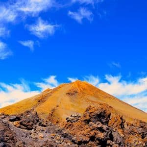 La cima arancione di una montagna vulcanica si erge sopra rocce laviche aspre sotto un cielo azzurro intenso con nuvole sottili.