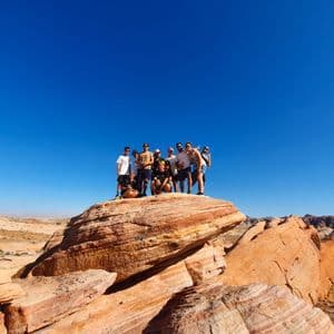 Eine WeRoad-Gruppe posiert gemeinsam auf einer großen roten Felsformation in einer Wüstenlandschaft unter klarem blauem Himmel.