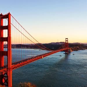 Eine große rote Hängebrücke, die eine Bucht bei Sonnenuntergang überspannt, mit Autos, die sie überqueren, und einer Stadtsilhouette im Hintergrund.