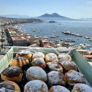 Una caja de pasteles variados con chocolate y azúcar glas se asoma a una ciudad costera con un gran puerto y una montaña a lo lejos.