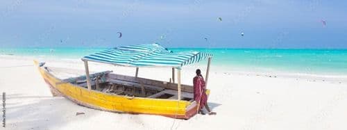 Une personne en robe rouge se tient à côté d'un bateau jaune avec un auvent rayé sur une plage de sable blanc, avec des kitesurfeurs sur l'océan turquoise.