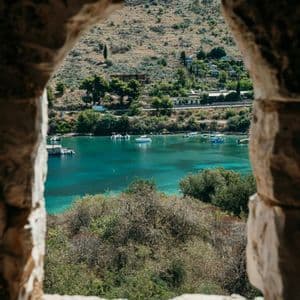 A view through a stone window overlooking a bay with turquoise water, small boats, and a green, hilly shoreline.