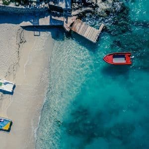 Top-down aerial view of a sandy beach with several boats on the sand and a red boat floating in turquoise water by a dock.