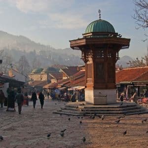Auf einem gepflasterten Platz steht ein verzierter Holzbrunnen mit einer grünen Kuppel, während Spaziergänger an Marktständen vorbeiflanieren und Tauben sich am Boden sammeln.