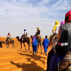 Un viaggio di gruppo WeRoad cavalca cammelli in fila attraverso vaste dune di sabbia arancioni sotto un cielo parzialmente nuvoloso.