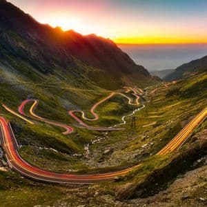 Lichtspuren von Autos beleuchten eine kurvenreiche Straße durch einen grasbewachsenen Gebirgspass, während die Sonne am Horizont untergeht.