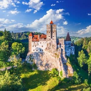 Un castello bianco con tetti di tegole rosse si erge su una scogliera rocciosa circondato da una fitta foresta verde sotto un cielo azzurro con nuvole.