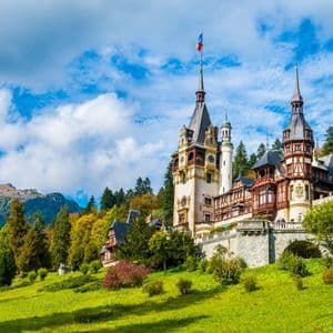 Ein großes, verziertes Schloss mit mehreren Türmen thront auf einem grünen, grasbewachsenen Hügel, der von einem dichten Wald und Bergen unter einem blauen, bewölkten Himmel umrahmt wird.