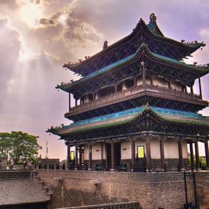 Una tradizionale pagoda cinese con tetti di tegole verdi si erge su un muro di pietra sotto un cielo con nuvole e raggi di sole penetranti.
