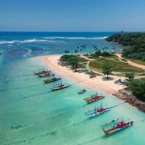 An aerial view of colorful fishing boats moored in a line in shallow, turquoise water next to a sandy beach.