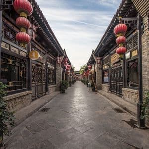 A narrow paved street flanked by traditional brick buildings decorated with red paper lanterns under a cloudy sky.
