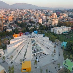 Une vue aérienne d'un bâtiment moderne, blanc, en forme de pyramide, avec des boîtes colorées sur son toit, situé dans une ville animée au coucher du soleil.