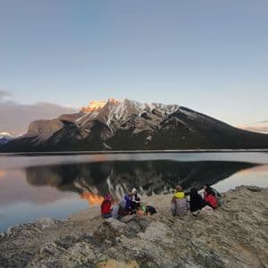 Un gruppo WeRoad su una riva rocciosa, ammira il tramonto che illumina una montagna innevata riflessa in un lago calmo.