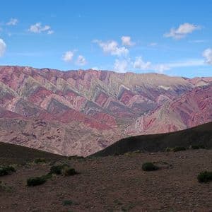Una cordillera con coloridas capas rayadas de rojo, morado y beige bajo un cielo azul parcialmente nublado.