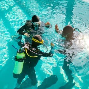 Un voyage en groupe WeRoad de trois personnes, équipées de matériel de plongée, apprenant des techniques de plongée dans une piscine ensoleillée.
