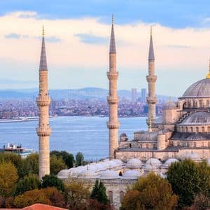 Una grande moschea con sei minareti e diverse cupole si affaccia su uno specchio d'acqua, con una nave da carico e uno skyline cittadino sullo sfondo.