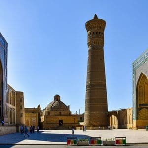 Un alto minarete de ladrillo y edificios ornamentados con arcos de azulejos azules rodeando una plaza de piedra iluminada por el sol bajo un cielo despejado.