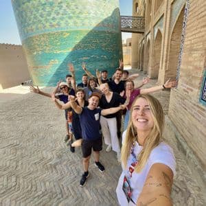 Un groupe WeRoad en voyage prend un selfie de groupe dans une rue pavée, à côté d'une grande tour carrelée turquoise.