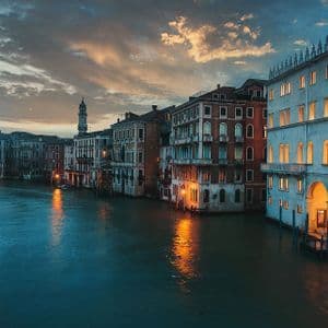 Edifici storici con finestre illuminate costeggiano un ampio canale al crepuscolo, le cui luci si riflettono sull'acqua.