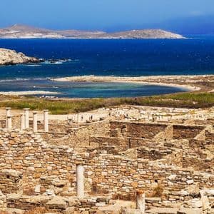 Antiche rovine in pietra con colonne bianche che si affacciano su un mare blu brillante e isole lontane sotto un cielo sereno.
