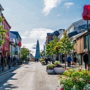 Eine sonnige Stadtstraße mit farbenfrohen Gebäuden und Bäumen, Menschen gehen auf einen hohen Kirchturm in der Ferne zu.