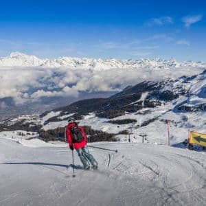 Ein Skifahrer in roter Jacke fährt einen schneebedeckten Berghang hinab, mit Blick auf ein Wolkenmeer und entfernte Gipfel unter einem klaren blauen Himmel.
