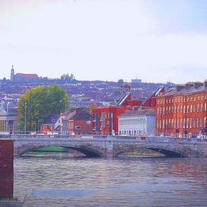 Un pont de pierre à plusieurs arches enjambe une large rivière, débouchant sur une ville aux bâtiments colorés et une colline en arrière-plan.